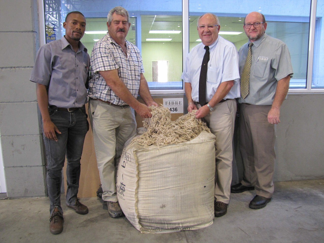 Four individuals standing behind a burlap sack filled with mohair cuttings, placed in front of a small wall sign featuring the words 