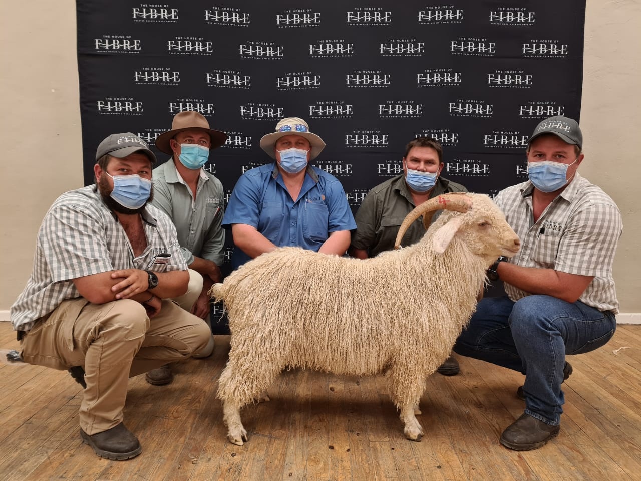 Five individuals wearing masks pose with an Angora goat in front of a backdrop with the logo of The House of Fibre.