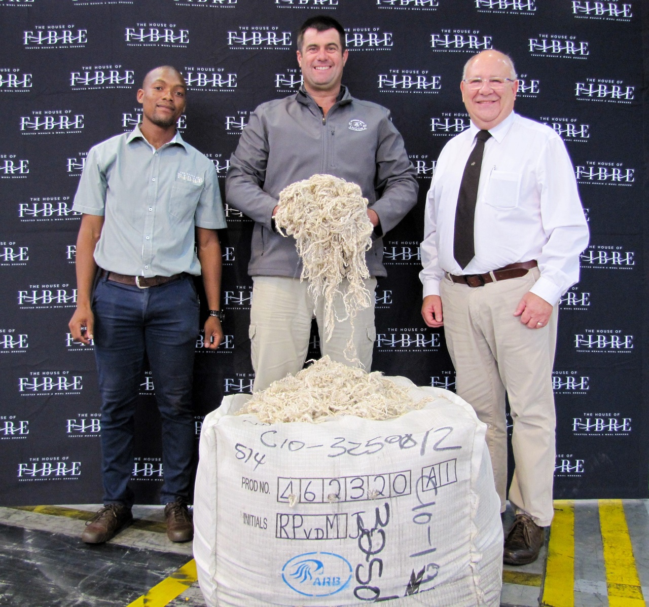 Three individuals standing behind a large bale of wool, with a backdrop featuring the logo 