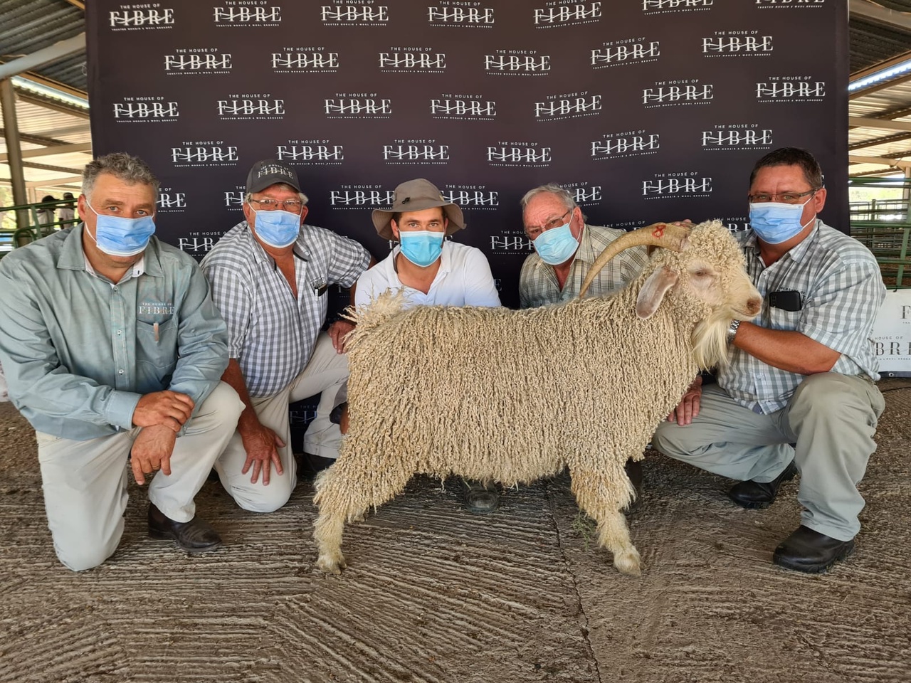 Five individuals wearing masks pose with a sheep in front of a banner with multiple House of Fibre logos. They are at the Van Hassalt Angora Auction 2021.