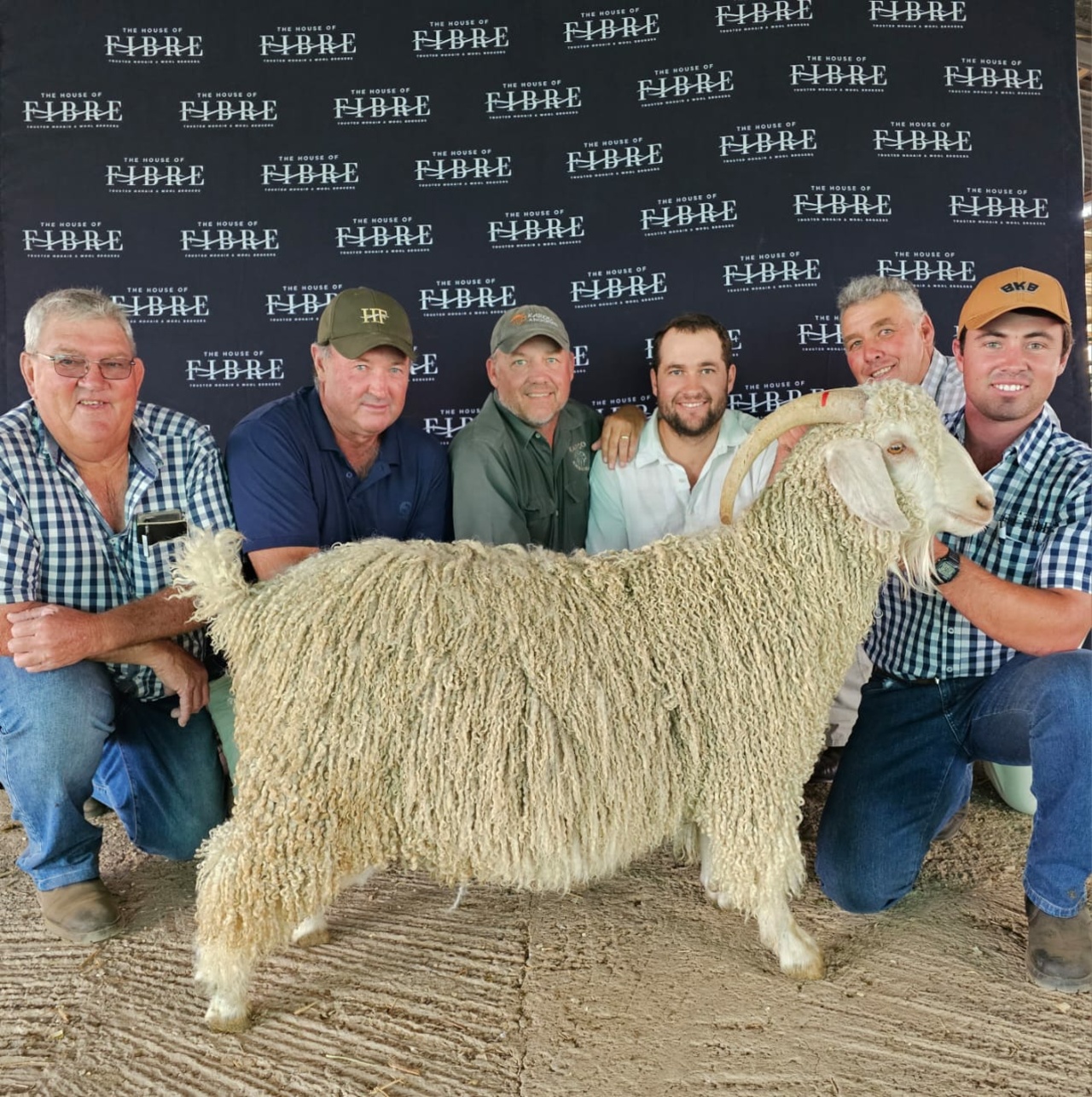 Group of six individuals posing with a large sheep at an event, standing in front of a backdrop with the logo of The House of Fibre.