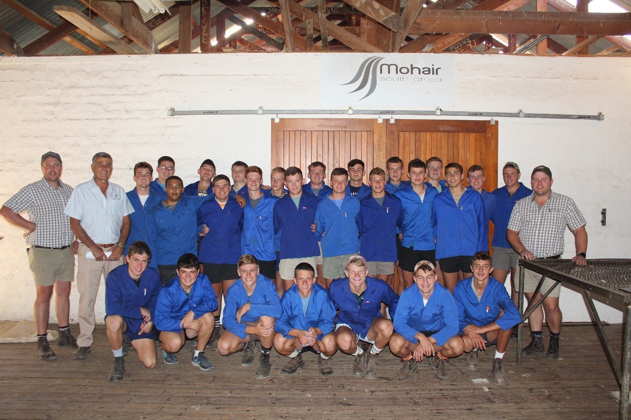 Group of individuals from Landbouskool Marlow, in blue shirts posing for a photo in front of a Mohair sign in a rustic indoor setting.