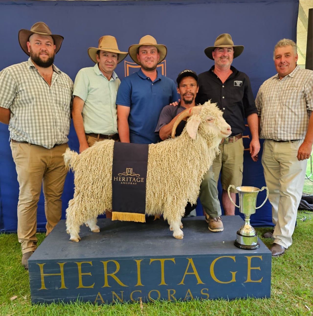 Group of five individuals posing with a champion angora goat at a livestock show, standing under a tent with a banner reading 